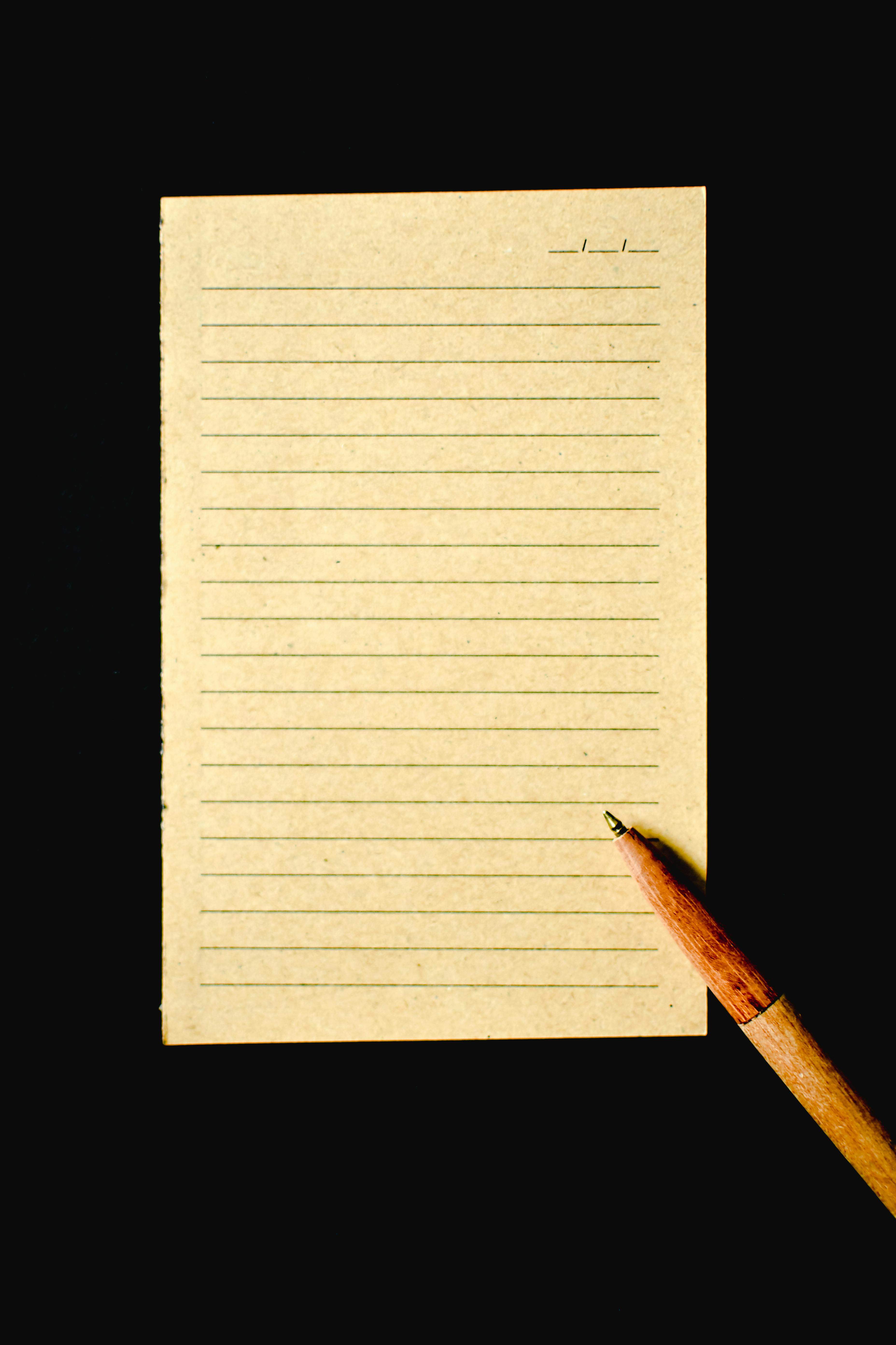 A blank lined notebook page with a wooden pen resting on it, lit against a black background